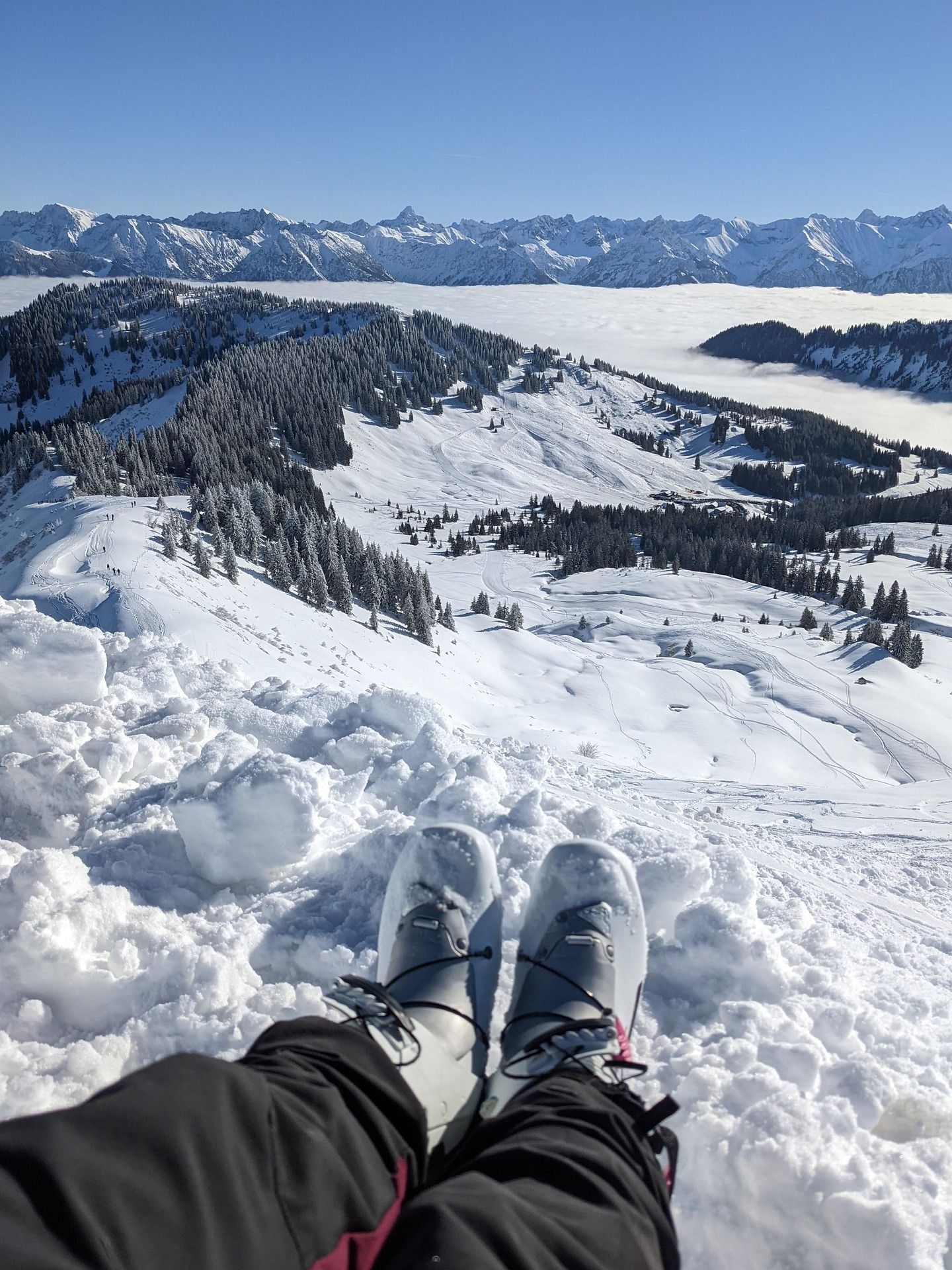 Vue à la première personne de chaussures de ski reposant dans la neige profonde, dominant une vaste chaîne de montagnes enneigée au-dessus d'une mer de nuages.
