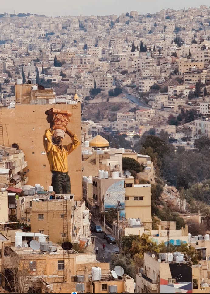 Un grande murale di un uomo che porta un capitello di colonna su un edificio domina un paesaggio urbano denso e collinare.