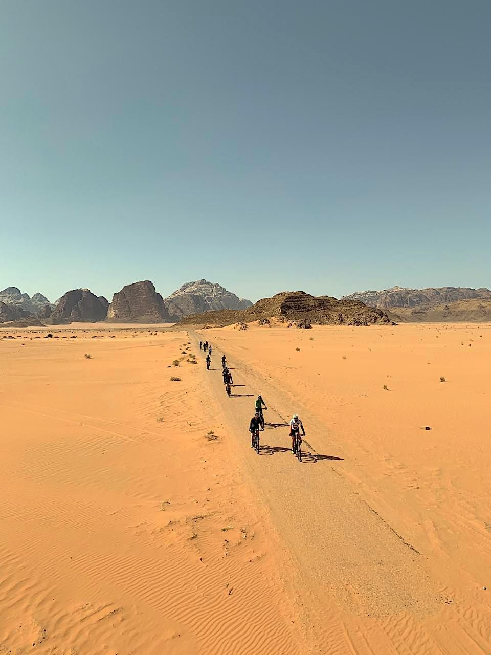 Un gruppo WeRoad in viaggio, pedala in bicicletta in fila su un sentiero sterrato che attraversa un vasto deserto con montagne rocciose in lontananza.