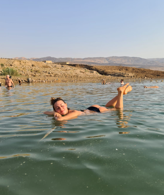 Una donna sorridente galleggia sulla schiena in acqua calma con i piedi sollevati, con una costa rocciosa e arida e colline sullo sfondo.