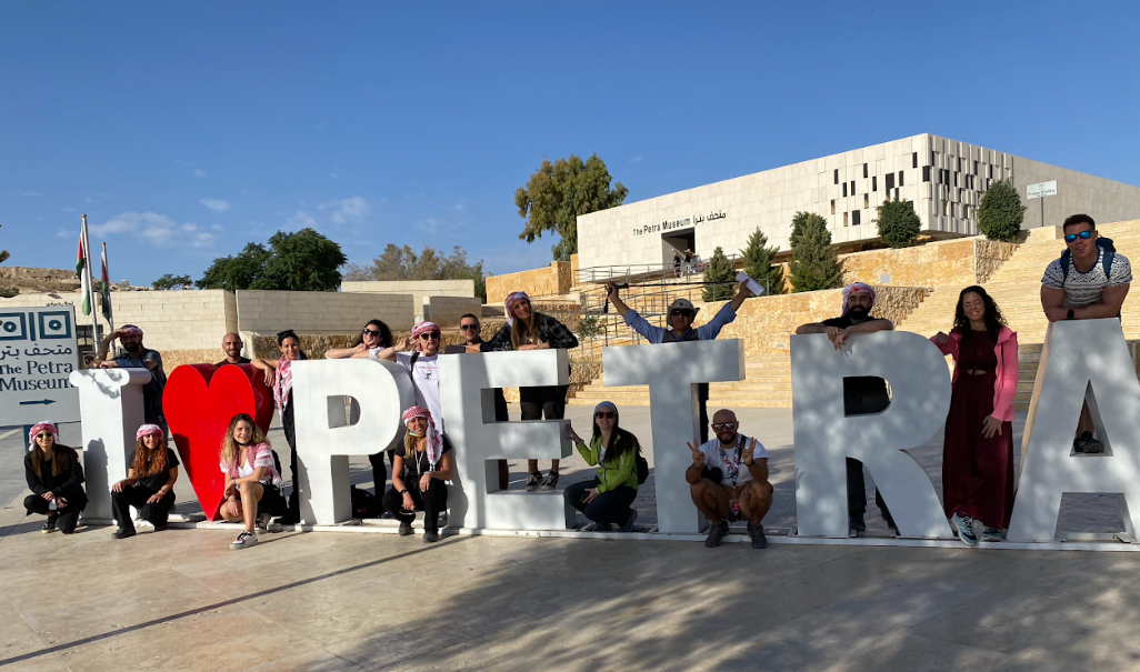 Un gruppo in viaggio WeRoad si mette in posa per una foto attorno alla grande insegna 'I ❤️ PETRA' fuori dal Museo di Petra, sotto un cielo sereno.