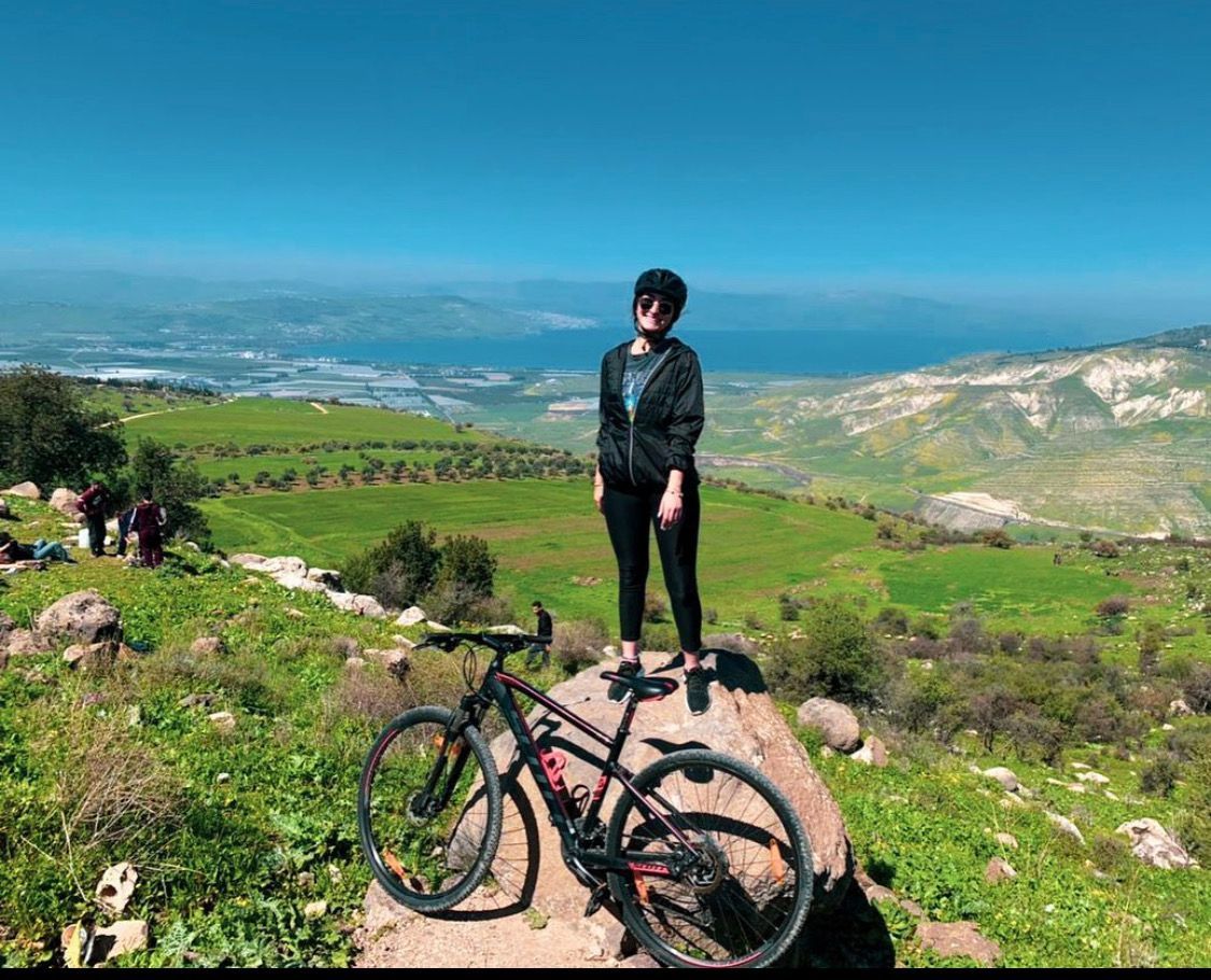 Una donna con l'abbigliamento da ciclismo è in piedi su una roccia accanto alla sua mountain bike, con vista su una valle verde e uno specchio d'acqua.