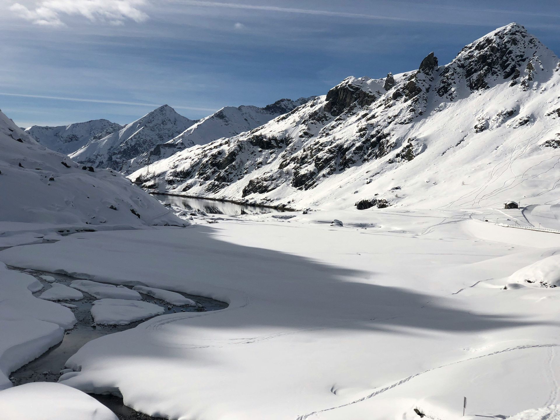 Un lago parzialmente ghiacciato si snoda attraverso una valle innevata circondata da cime montuose illuminate dal sole sotto un cielo blu.