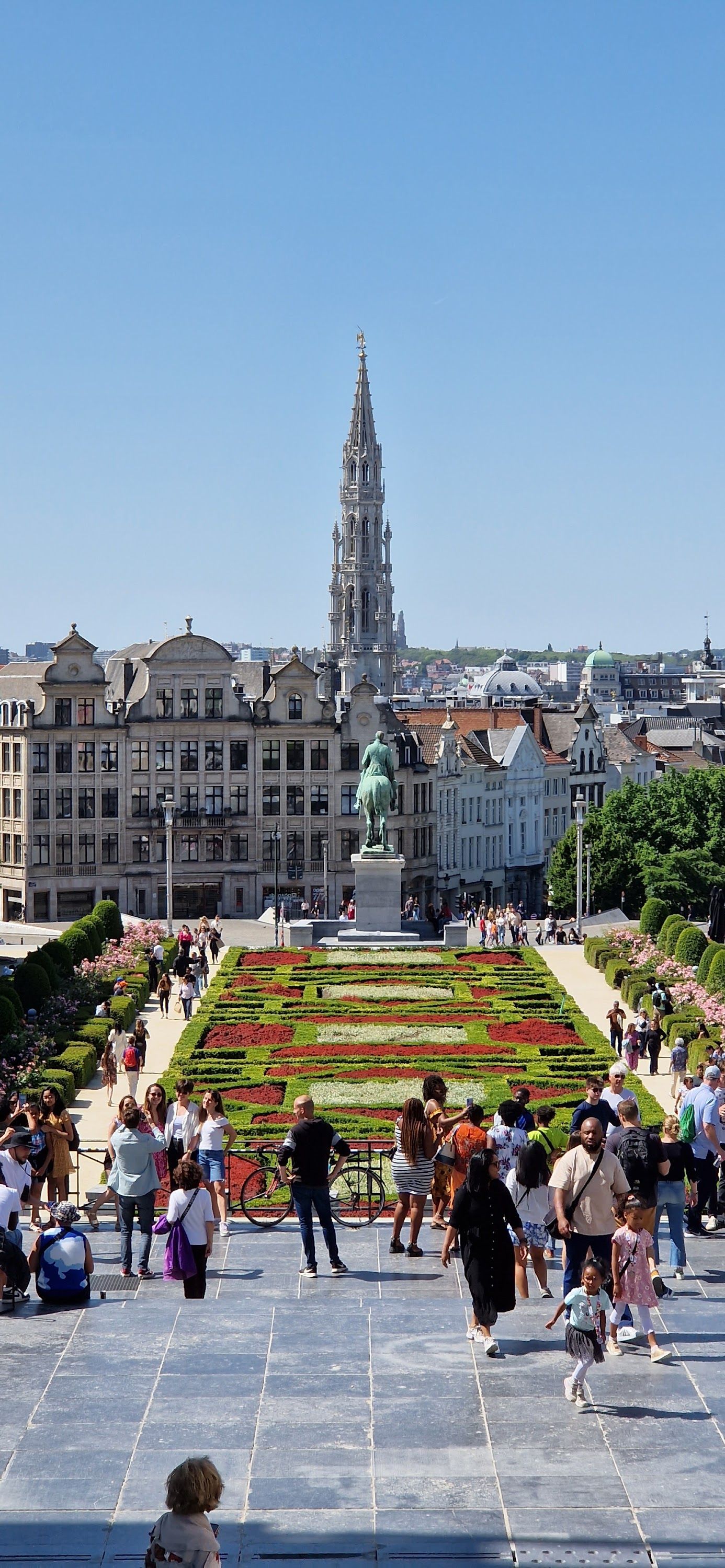 Des gens se promènent dans un jardin formel sur une place de ville, avec des bâtiments historiques et une haute flèche en arrière-plan.