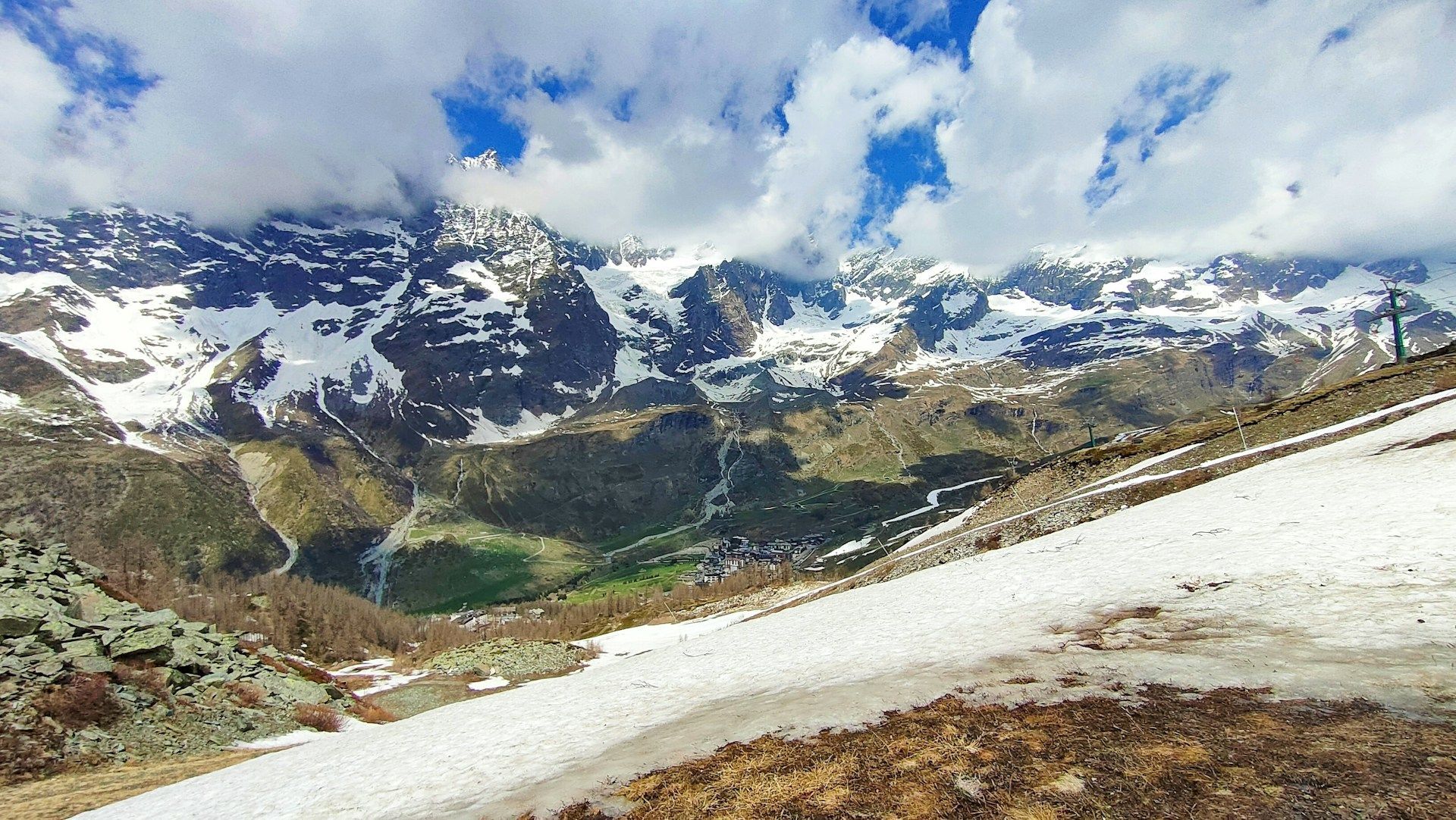 A panoramic view of a snow-capped mountain range over a green valley with a small village, seen from a snowy slope under a cloudy sky.