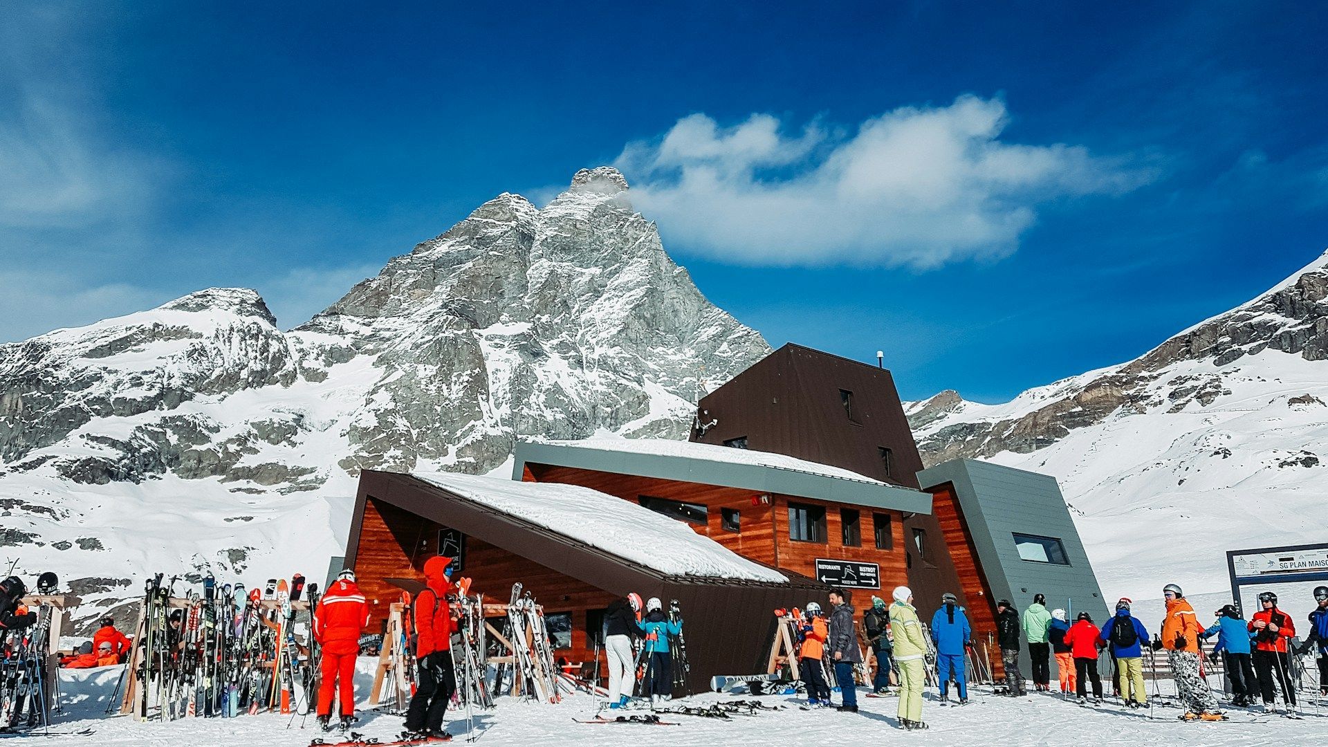 A WeRoad group trip with skis and snowboards gathers in front of a mountain lodge, with a large snow-covered peak under a blue sky.