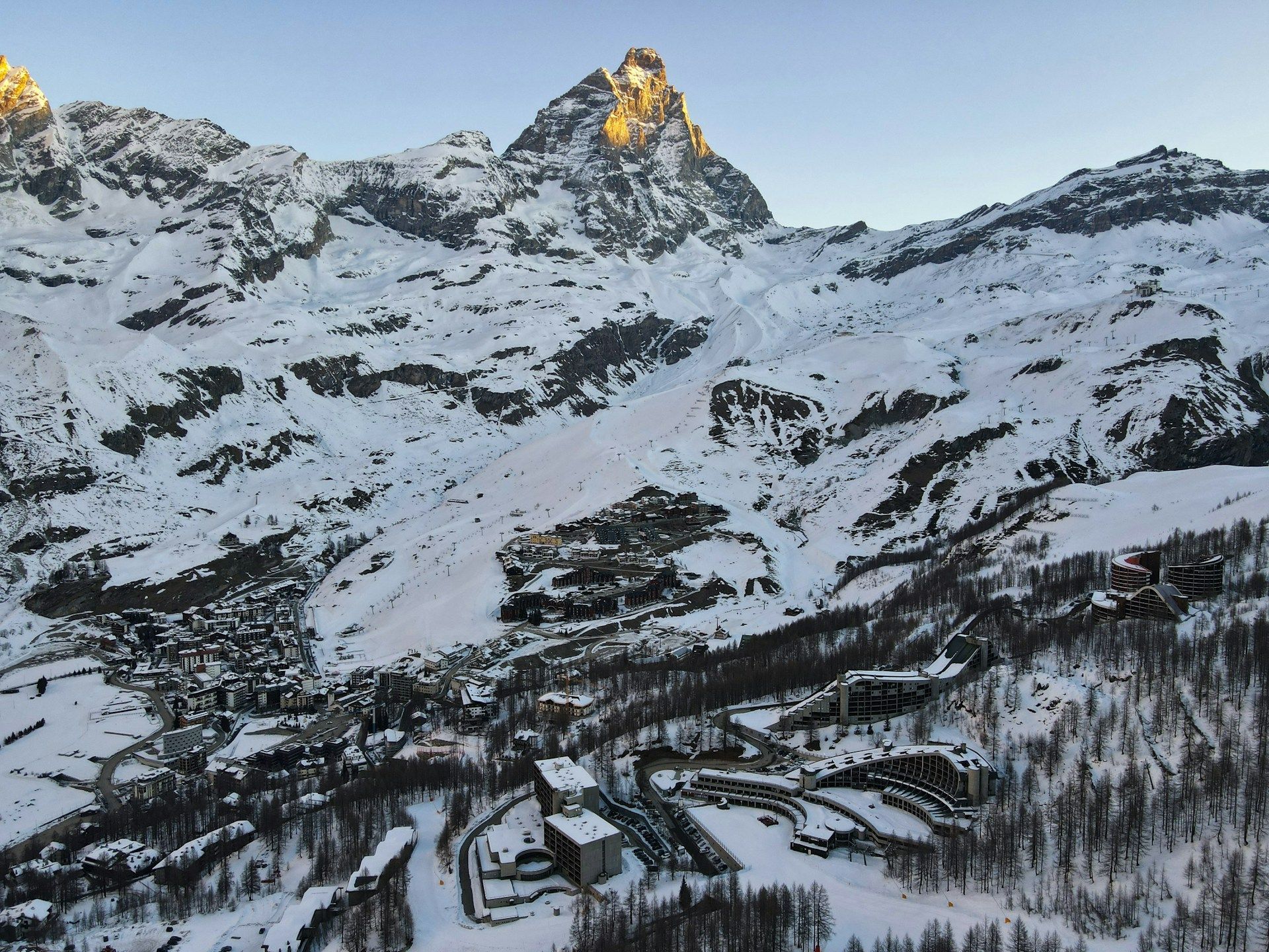 An aerial view of a ski village nestled in a snow-covered valley at the foot of a large, sunlit mountain peak.