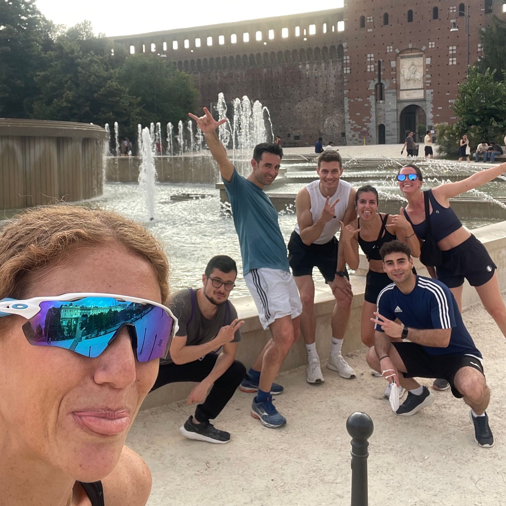A WeRoad group trip in athletic wear posing for a selfie in front of a large fountain and a historic brick castle.
