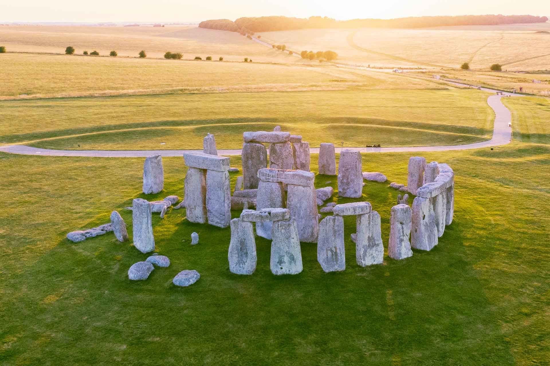 Una vista aerea del monumento di Stonehenge su un prato verde, illuminato dalla luce dorata del tramonto su campi ondulati.