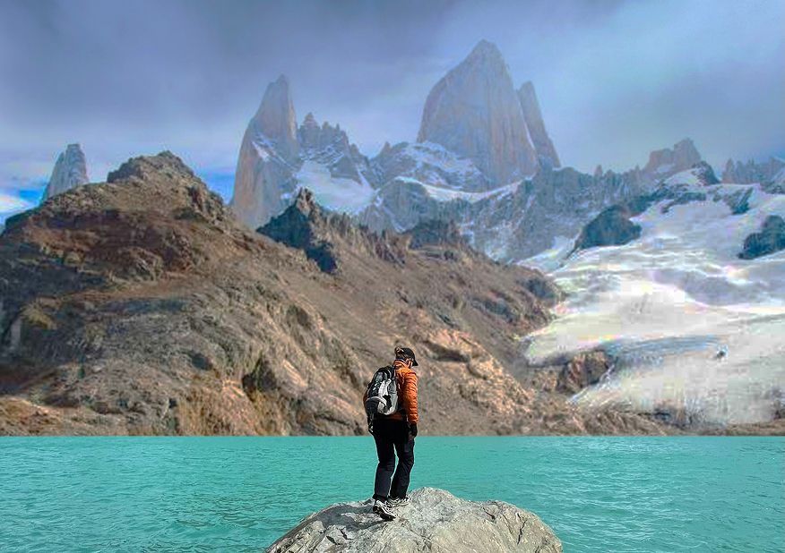 Un excursionista con mochila de pie sobre una roca con vistas a un lago turquesa y una cordillera irregular y nevada al fondo.