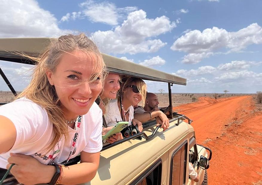 Des participants à un voyage de groupe WeRoad sourient pour un selfie depuis une jeep de safari décapotable, roulant sur une piste de terre rouge à travers une savane.