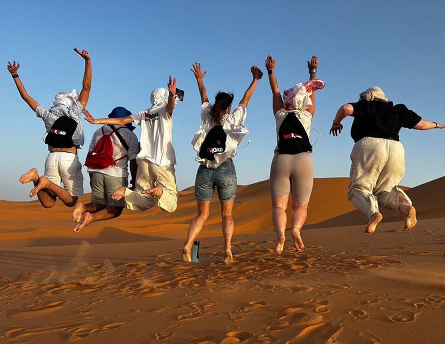 A WeRoad group trip seen from behind, jumping together on a vast desert sand dune under a clear sky.