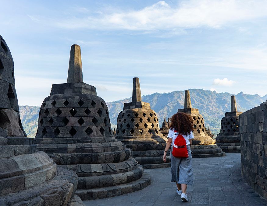 A woman with a red WeRoad backpack walks on a stone path among bell-shaped stupas, with mountains in the background.