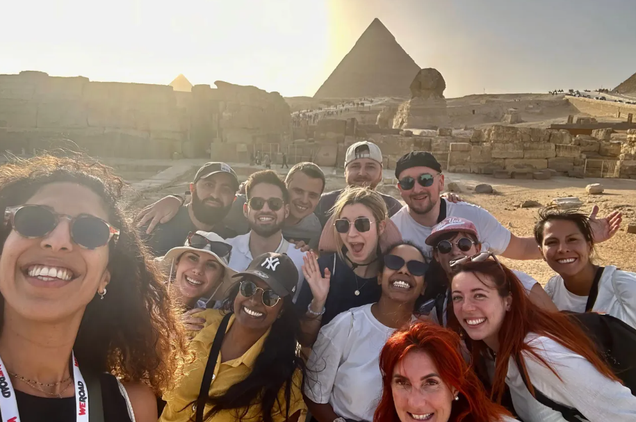 Un voyage de groupe WeRoad prend un selfie souriant avec une pyramide et le Sphinx en arrière-plan au coucher du soleil.