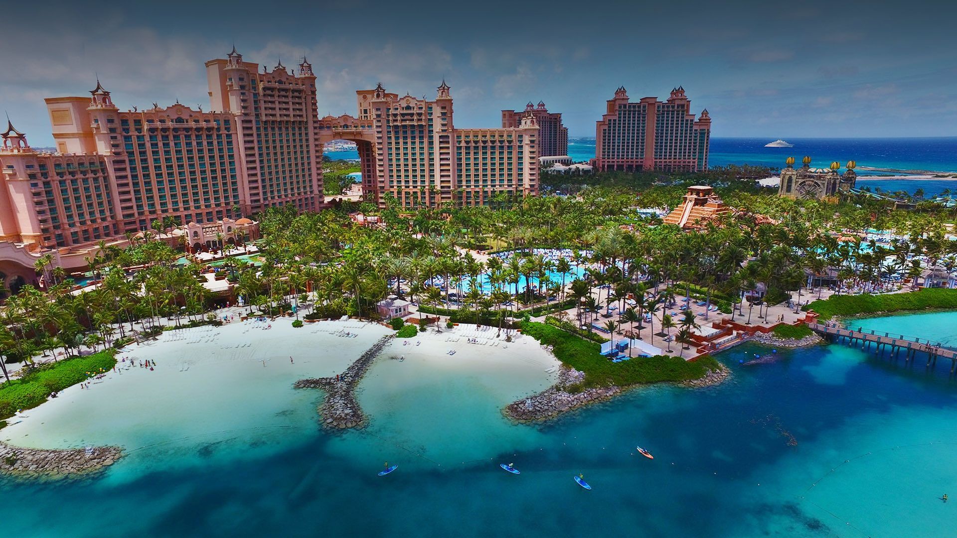 Vista aerea di un grande resort alberghiero rosa su una costa tropicale con spiagge di sabbia bianca, palme e acqua turchese.