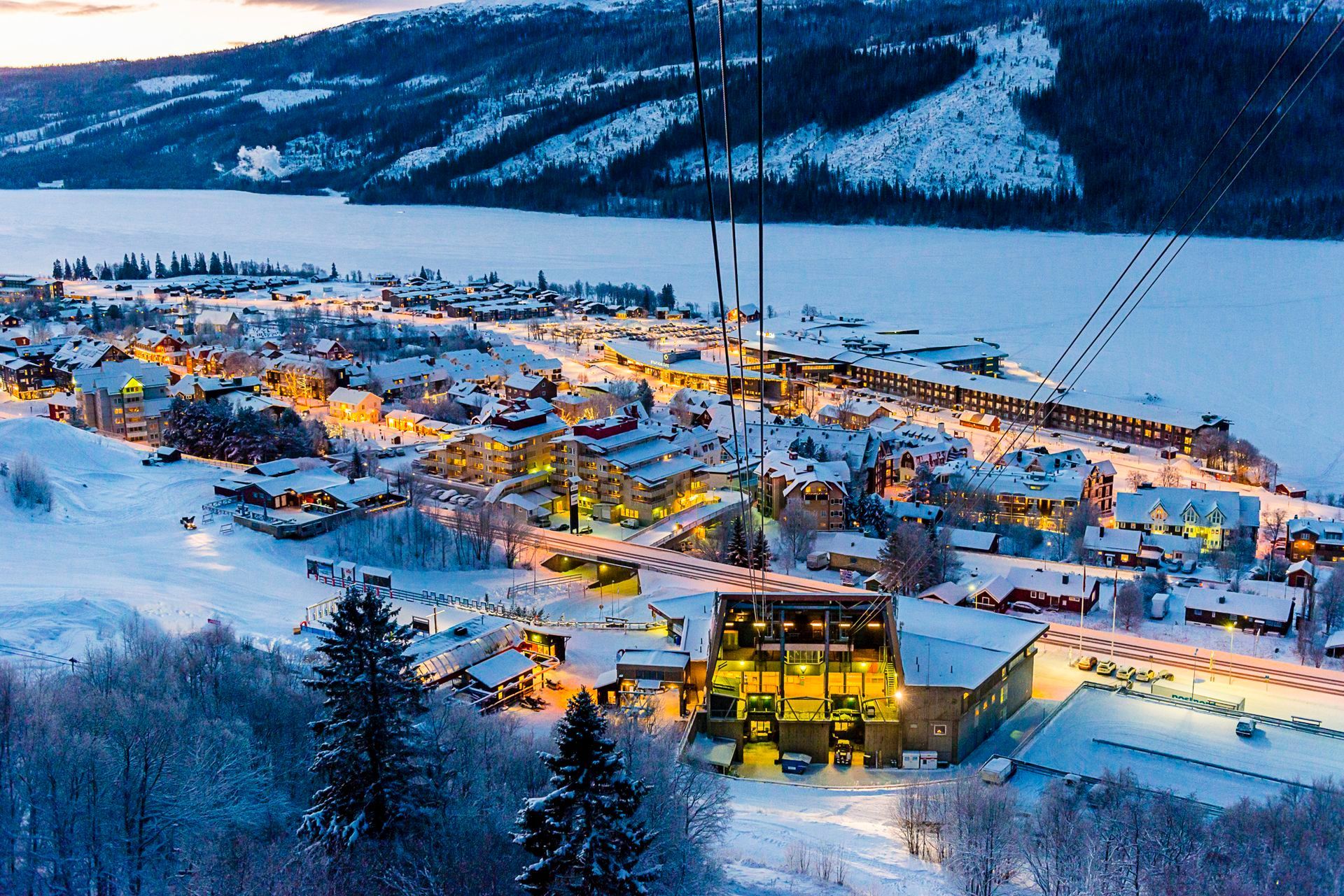 Una vista dall'alto di un villaggio innevato illuminato al crepuscolo, con un lago ghiacciato e montagne sullo sfondo.
