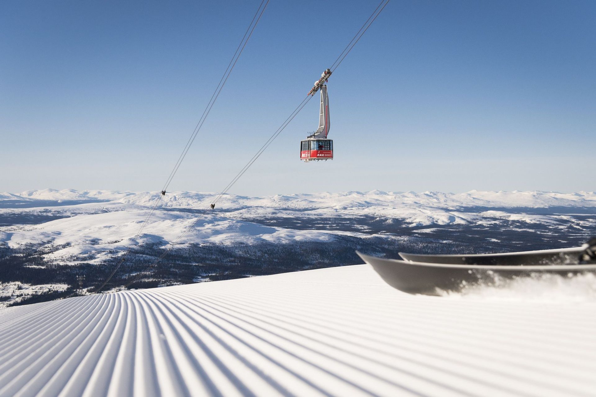 Una funivia viaggia sopra una pista da sci appena battuta, con le punte di uno snowboard in primo piano e montagne innevate sullo sfondo.