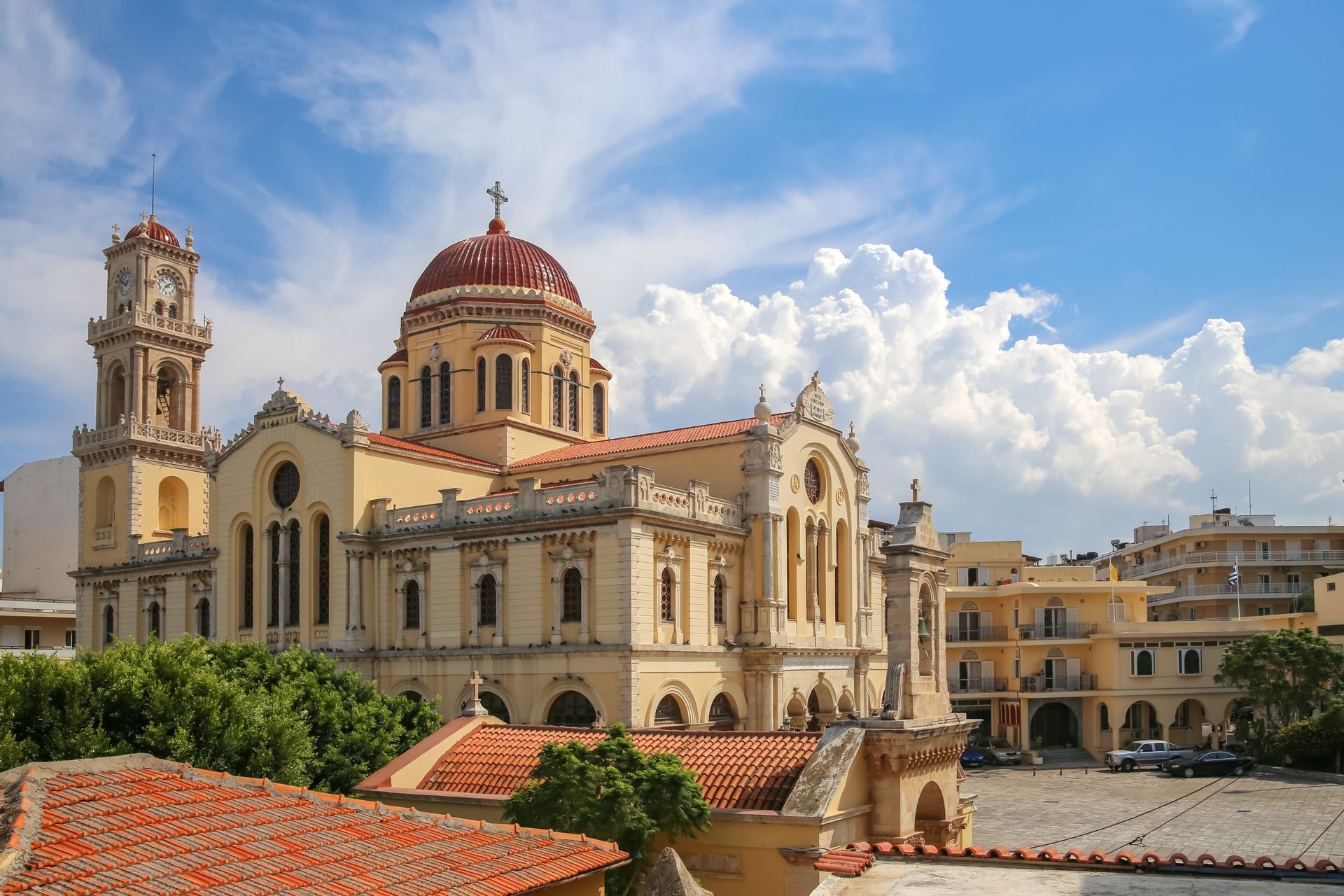 Una grande cattedrale gialla con una cupola rossa e una torre dell'orologio si erge in una piazza cittadina sotto un cielo azzurro nuvoloso.