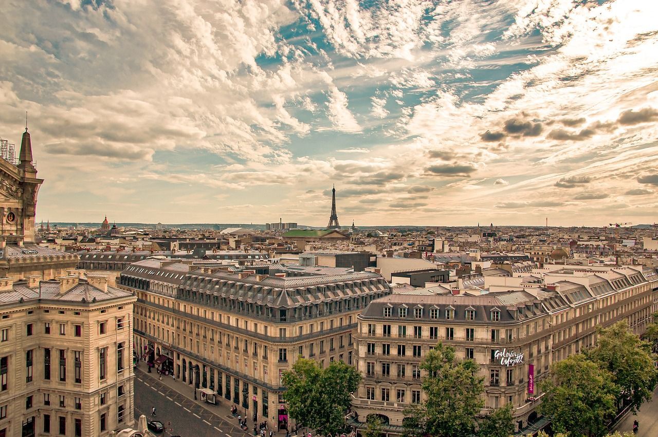 Une vue panoramique et plongeante des toits de Paris, avec la Tour Eiffel visible au loin sous un ciel parsemé de nuages.