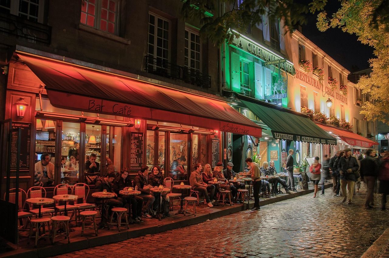 Des gens sont assis aux tables extérieures d'un bar-café, dînant dans une rue pavée la nuit sous des lumières chaudes rouges et vertes.