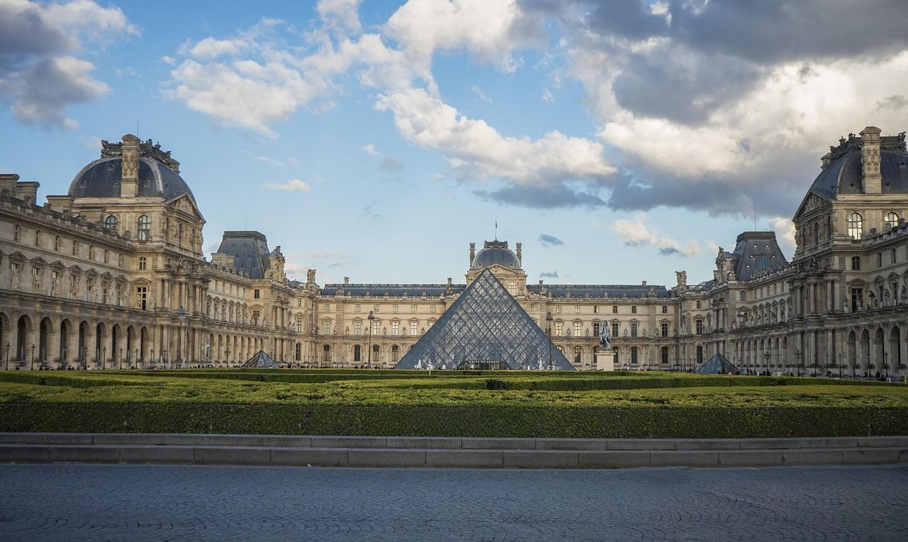 Une grande pyramide de verre se dresse dans la cour d'un bâtiment historique orné, avec une pelouse verte bien entretenue au premier plan sous un ciel nuageux.