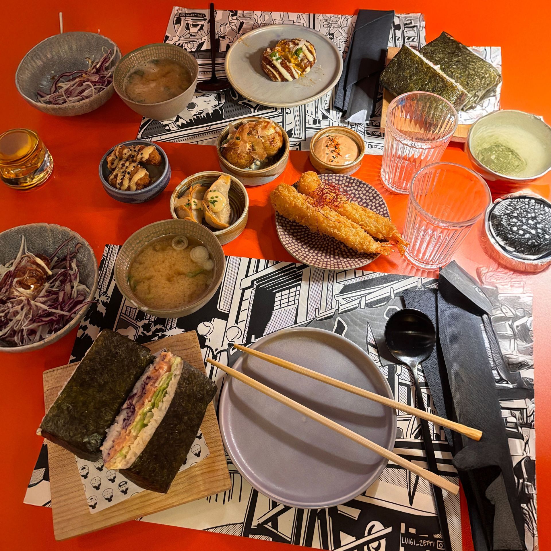 Una vista cenital de una variedad de platos japoneses como tempura, onigirazu y sopa miso, dispuestos sobre una mesa naranja vibrante.