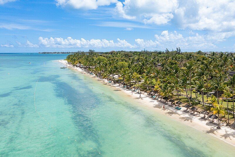Vista aerea di una spiaggia tropicale con acqua turchese, sabbia bianca e palme sotto un cielo azzurro e nuvoloso.