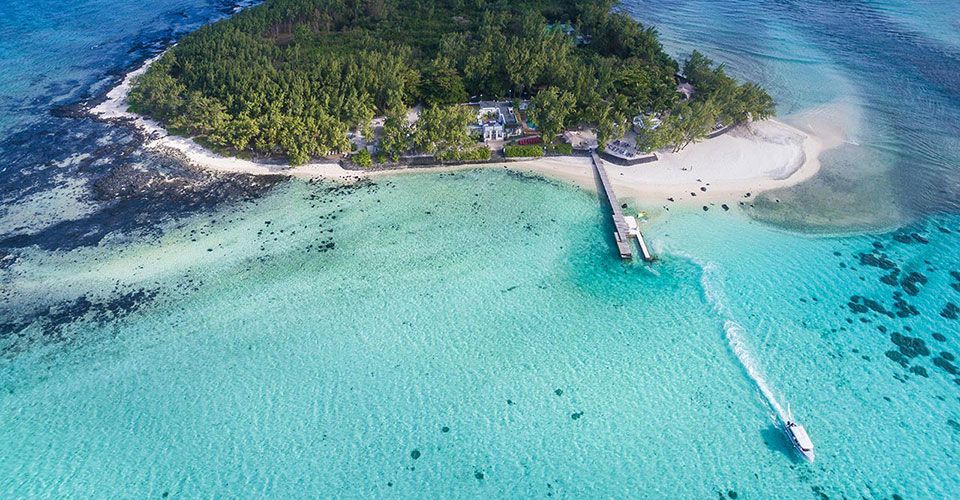 Una vista aerea di una piccola isola ricoperta di alberi, con una spiaggia sabbiosa e un molo, circondata da acqua cristallina turchese con un motoscafo che la attraversa.