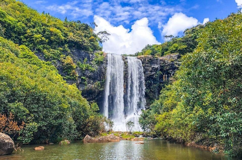 Una cascata si getta da una scogliera rocciosa in una pozza, incorniciata da fitti alberi verdi sotto un cielo azzurro parzialmente nuvoloso.