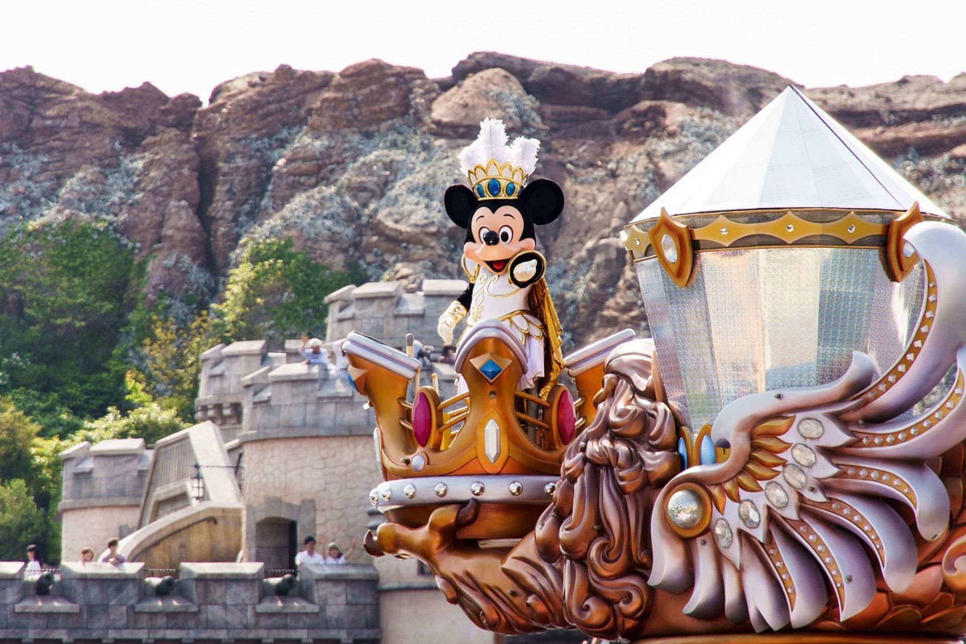 Mickey Mouse, vêtu d'un costume royal blanc et or, salue depuis un char de parade orné, avec un château et des montagnes en arrière-plan.