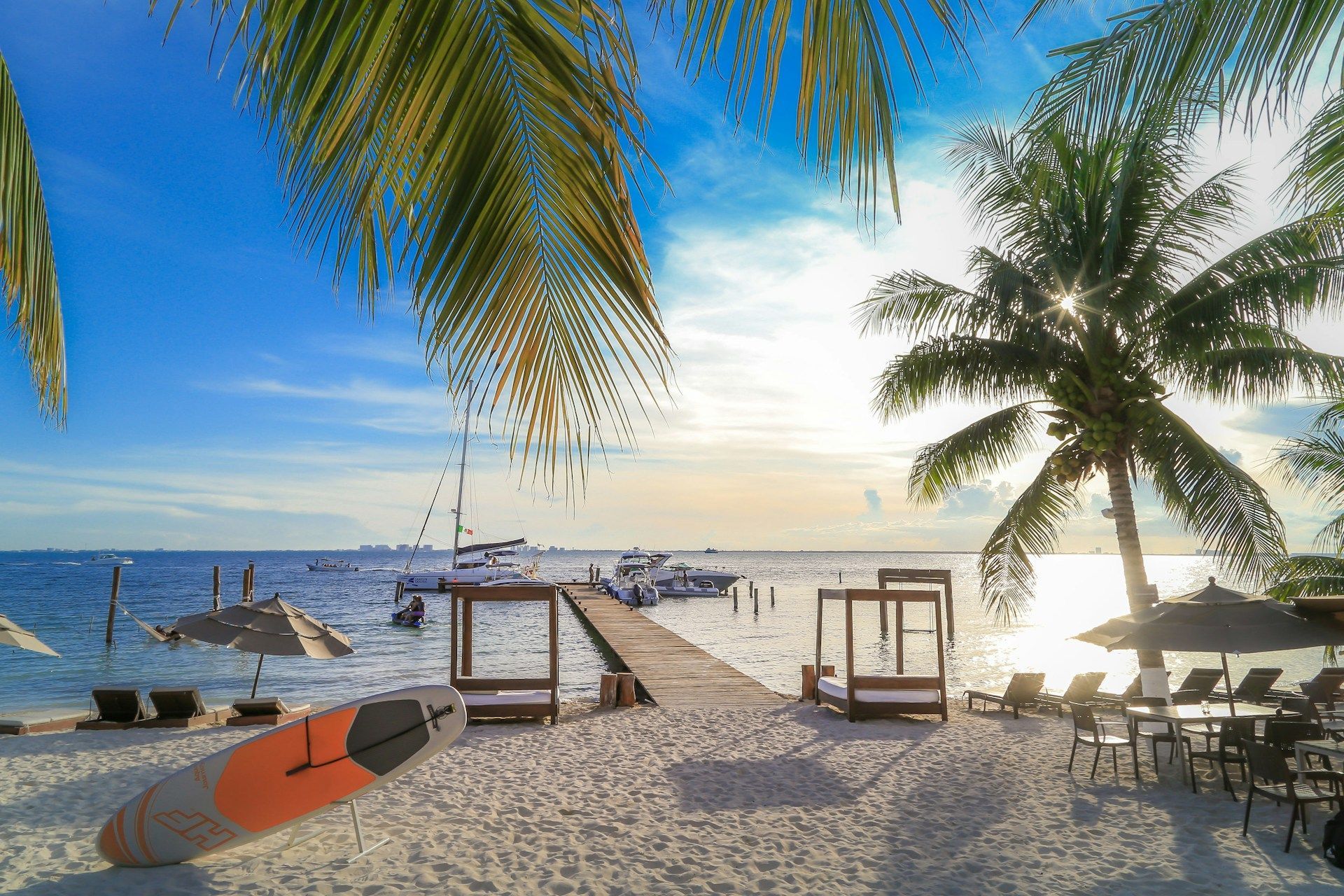 A wooden pier on a tropical beach with palm trees, lounge beds, and a paddleboard extending into the ocean at sunset.