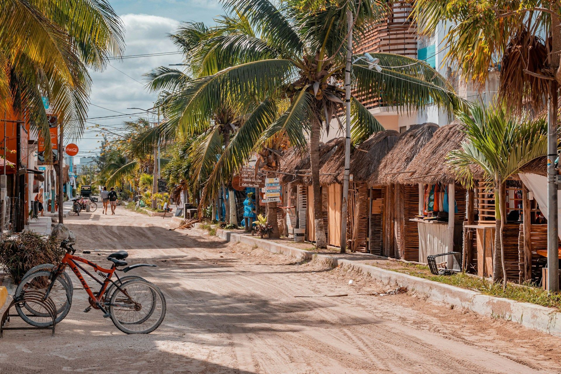 A red bicycle is parked on a sandy street lined with palm trees and rustic, thatched-roof shops.