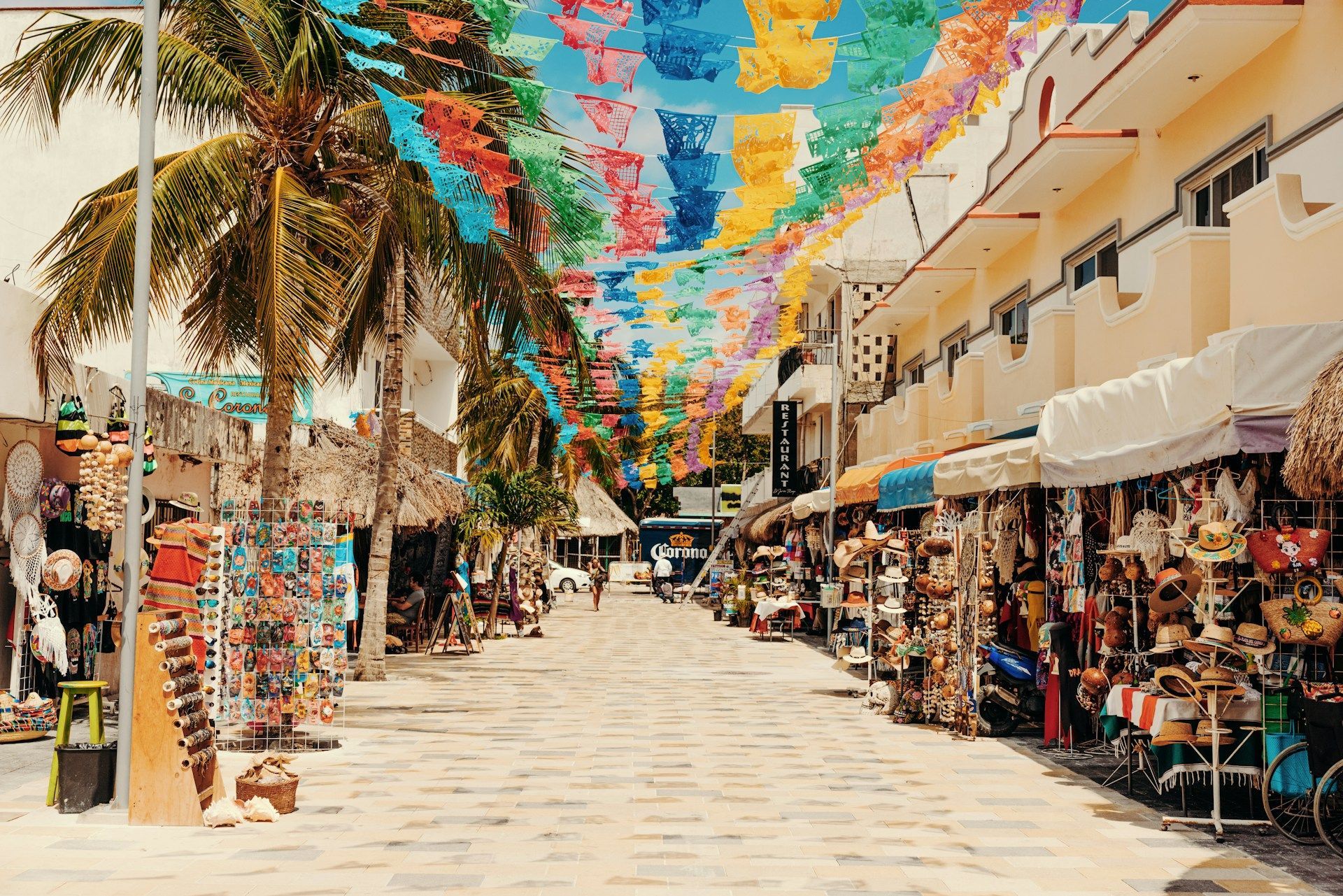 Una strada lastricata fiancheggiata da negozi e palme è decorata con bandiere di carta colorate appese in alto.