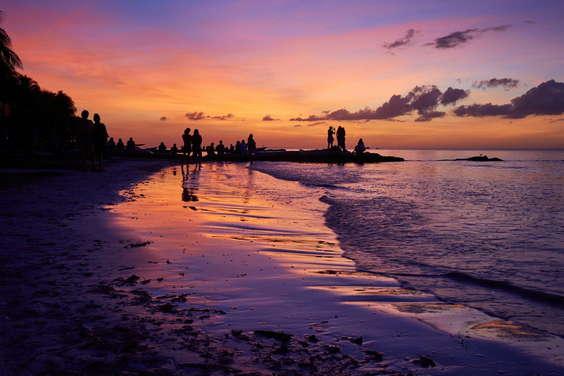 Sagome di un viaggio di gruppo WeRoad su una spiaggia durante un tramonto vibrante, con cieli viola e arancioni che si riflettono sulla sabbia bagnata.