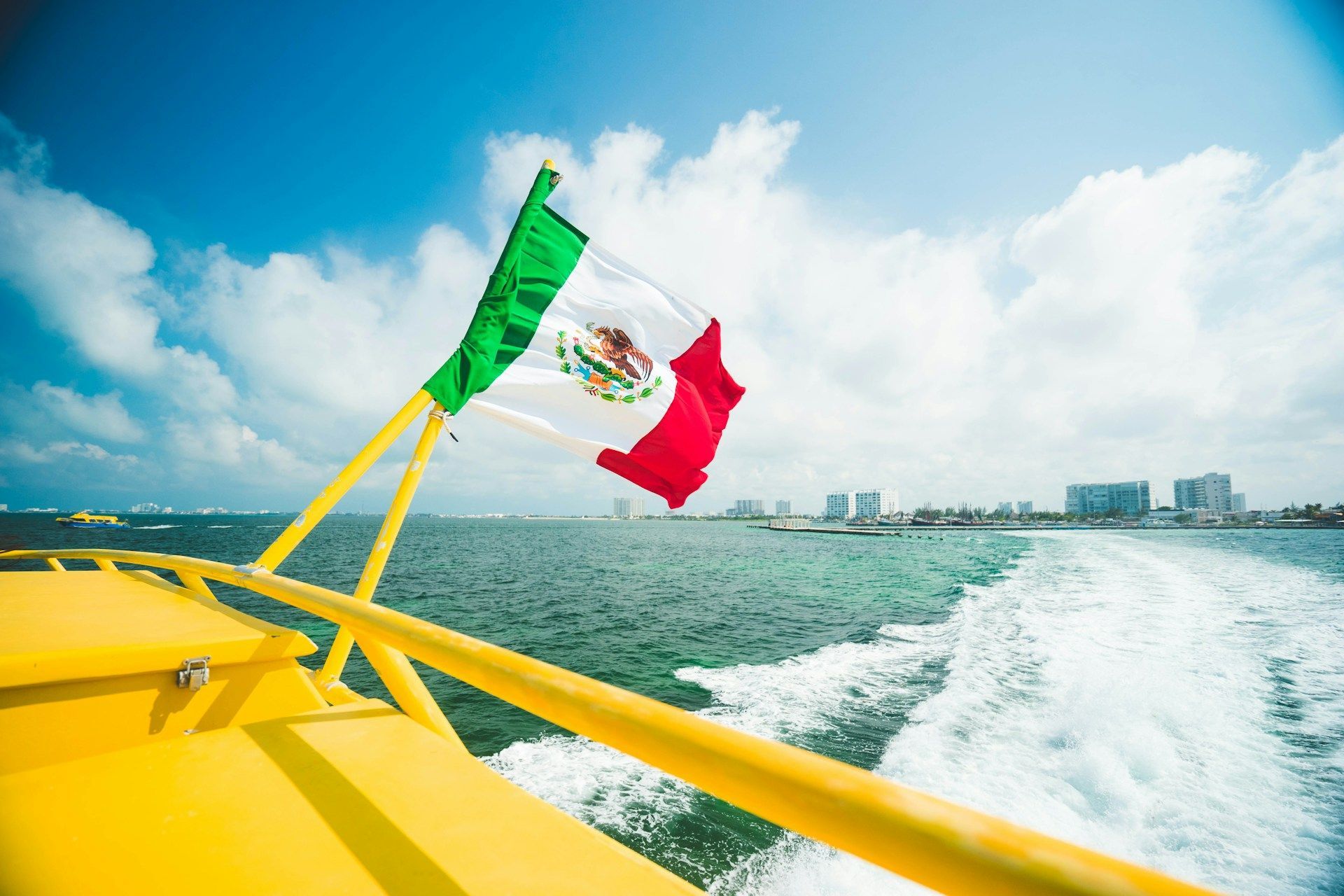 Una bandiera messicana sventola al vento sulla poppa di una barca gialla, creando una scia nell'acqua con lo skyline di una città sullo sfondo.
