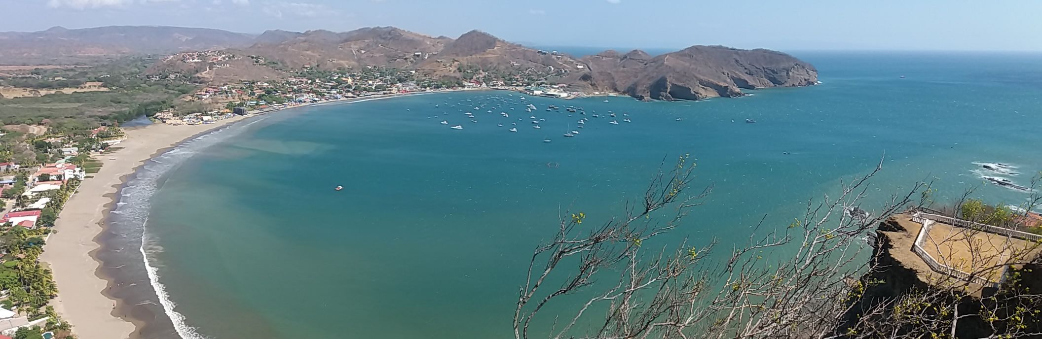 Ein Panoramablick auf eine große blaue Bucht mit vielen Booten, einem Sandstrand und einer Küstenstadt, die sich an trockene Hügel schmiegt.