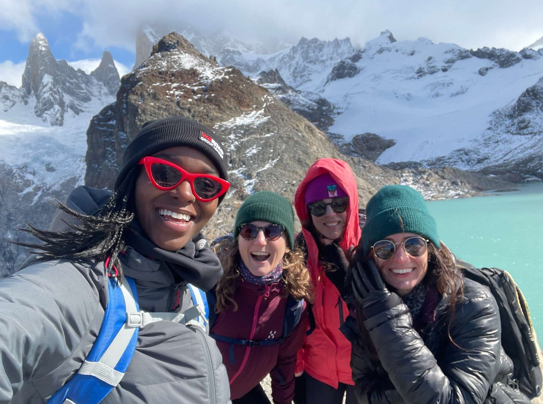 A WeRoad group trip of four women smiling for a selfie in front of a turquoise lake and snow-capped mountains.