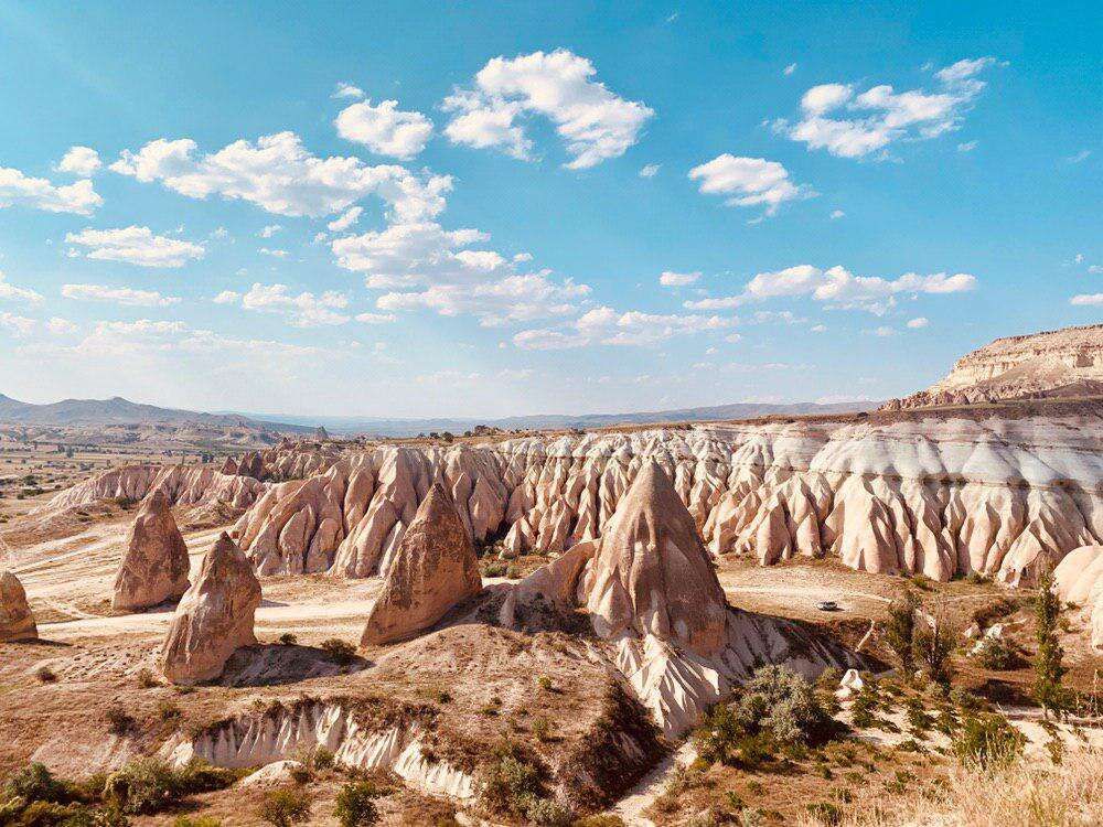 A wide landscape of conical rock formations in a dry valley under a bright blue sky with clouds.