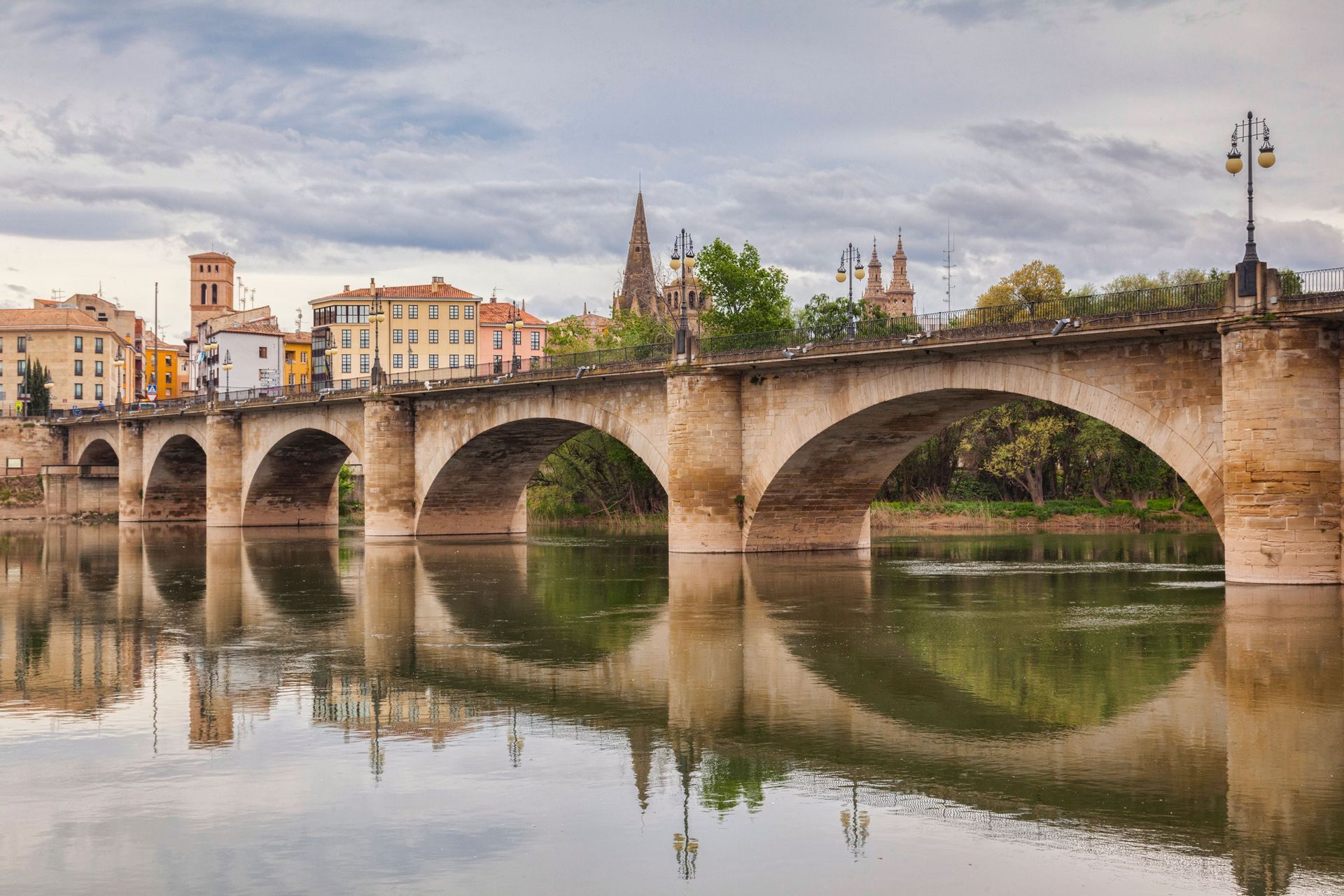 Un puente de piedra con múltiples arcos cruza un río tranquilo, reflejando el paisaje urbano histórico y el cielo nublado en su superficie.
