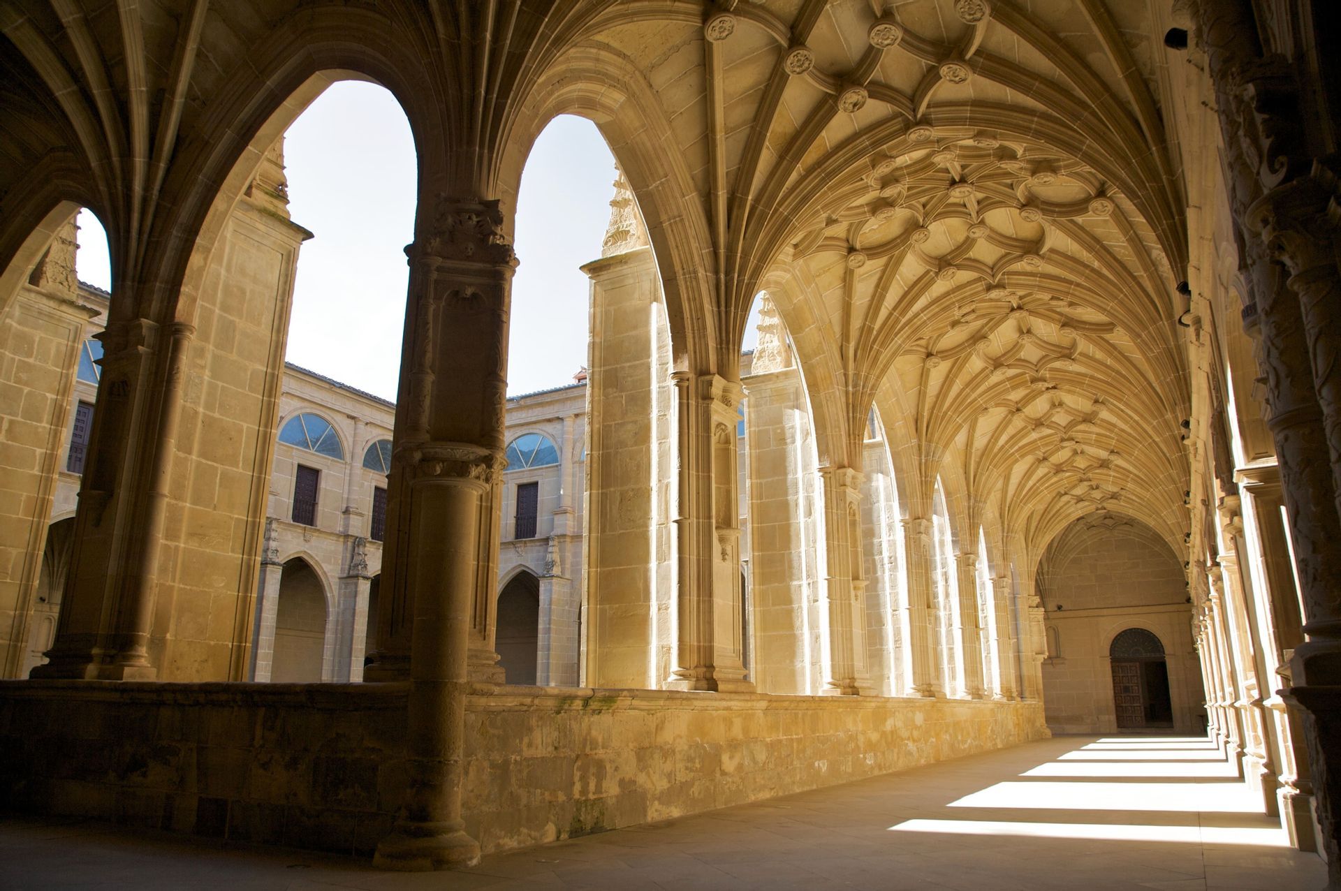 La luz del sol se filtra a través de los arcos góticos de un claustro de piedra, proyectando largas sombras por el pasillo con su ornamentado techo abovedado.