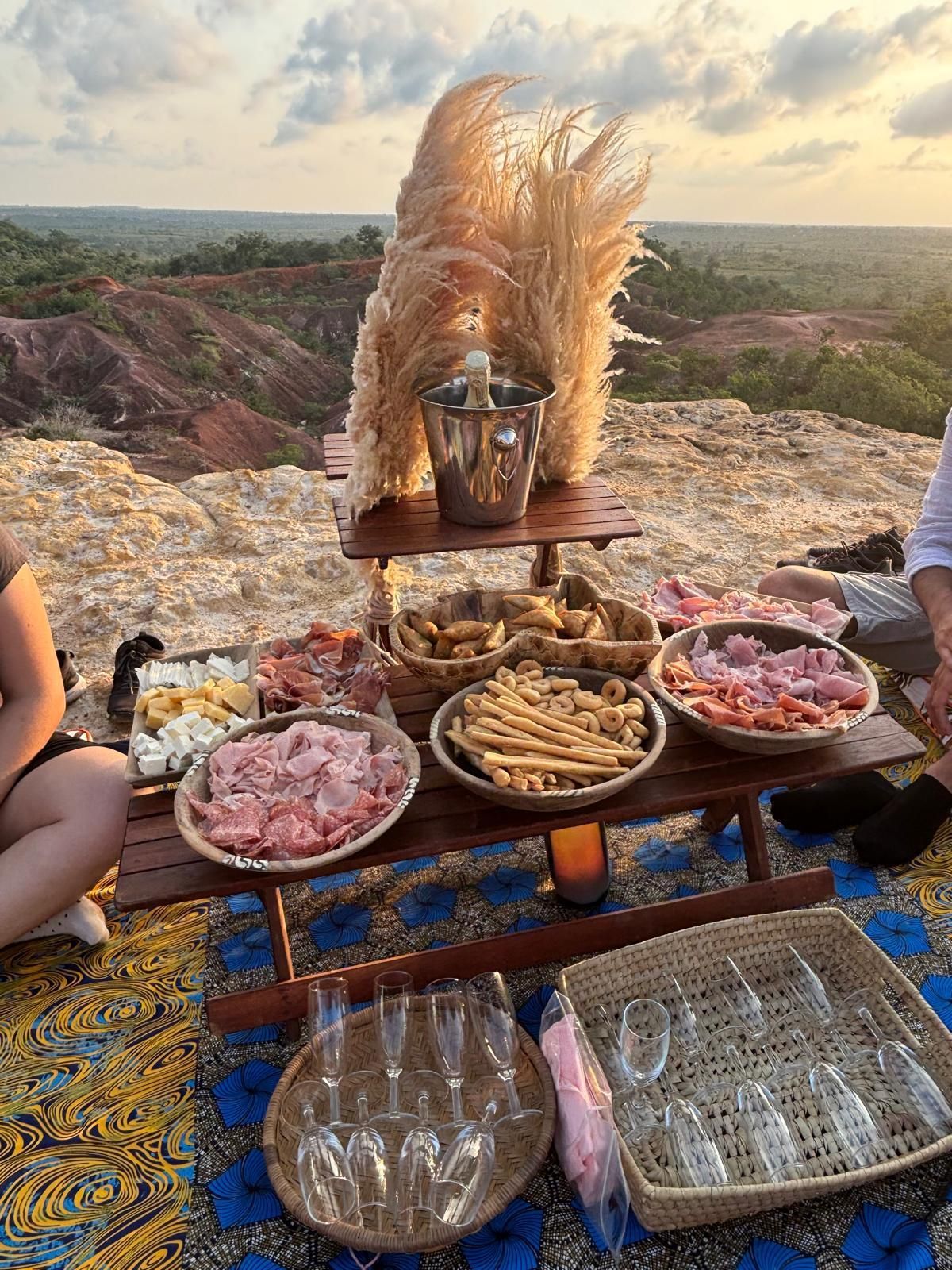 An outdoor picnic spread with sparkling wine, charcuterie, and cheese on a wooden table overlooking a canyon at sunset.
