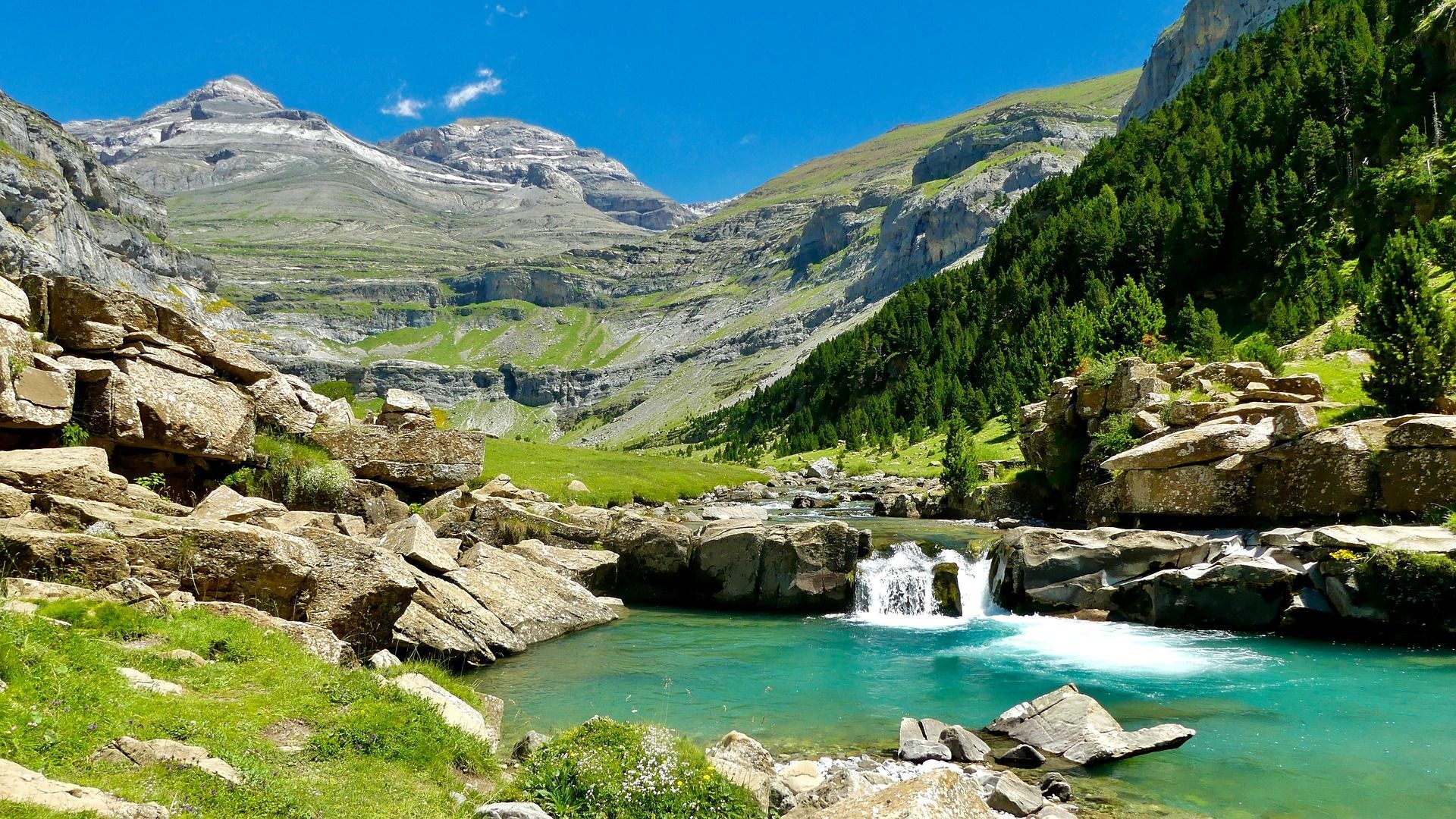 Un río turquesa con una pequeña cascada fluye a través de un valle de montaña verde bajo un cielo azul claro.