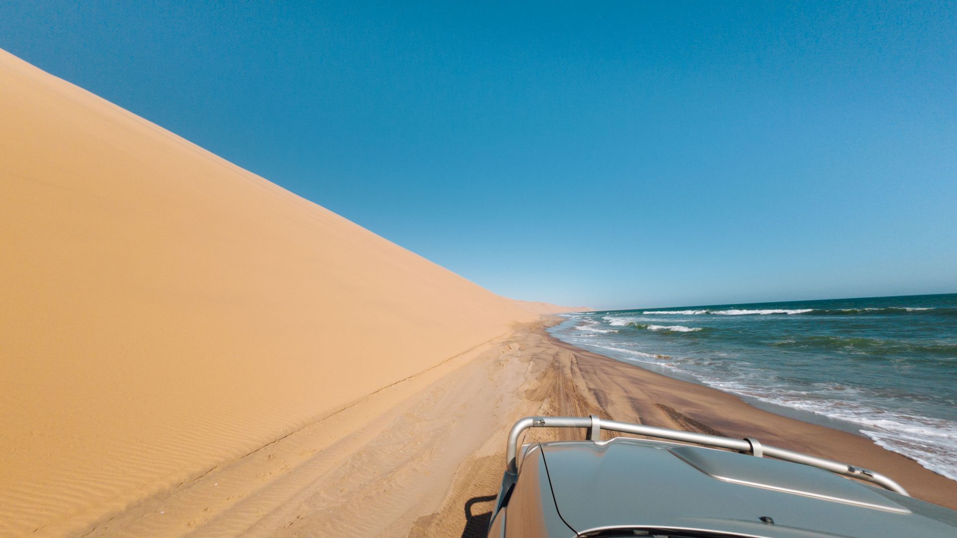 Una vista da un fuoristrada 4x4 che guida su una pista sabbiosa tra una grande duna di sabbia e le onde dell'oceano sotto un cielo limpido.