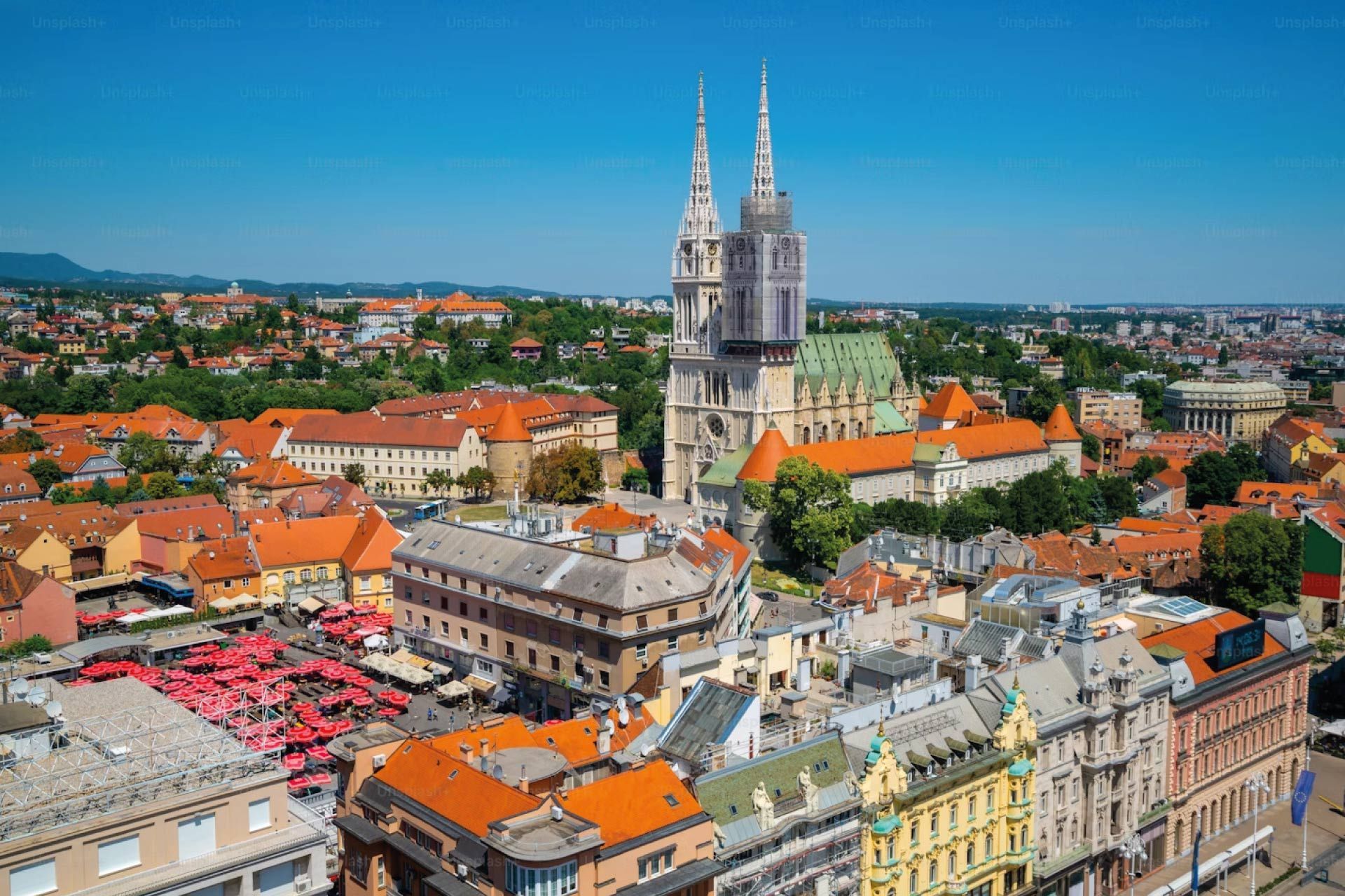 Une vue aérienne d'une ville historique aux toits de tuiles rouges, centrée sur une grande cathédrale à deux flèches sous un ciel bleu clair.