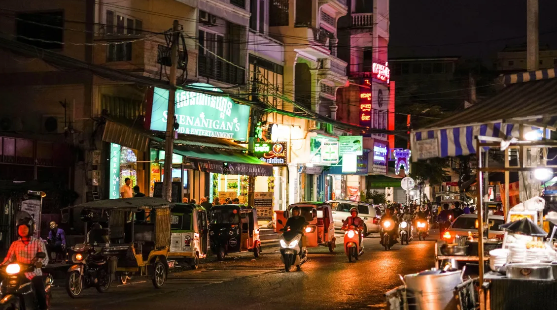 A busy city street at night filled with motorcycles, tuk-tuks, and illuminated by colorful neon signs on the buildings.