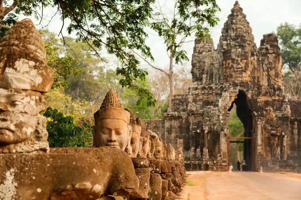 A row of weathered stone statues with carved faces lines a path leading to a large, ancient stone gate in a forest setting.