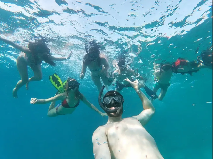An underwater selfie of a WeRoad group trip snorkeling together in clear blue water, with bubbles rising to the surface.