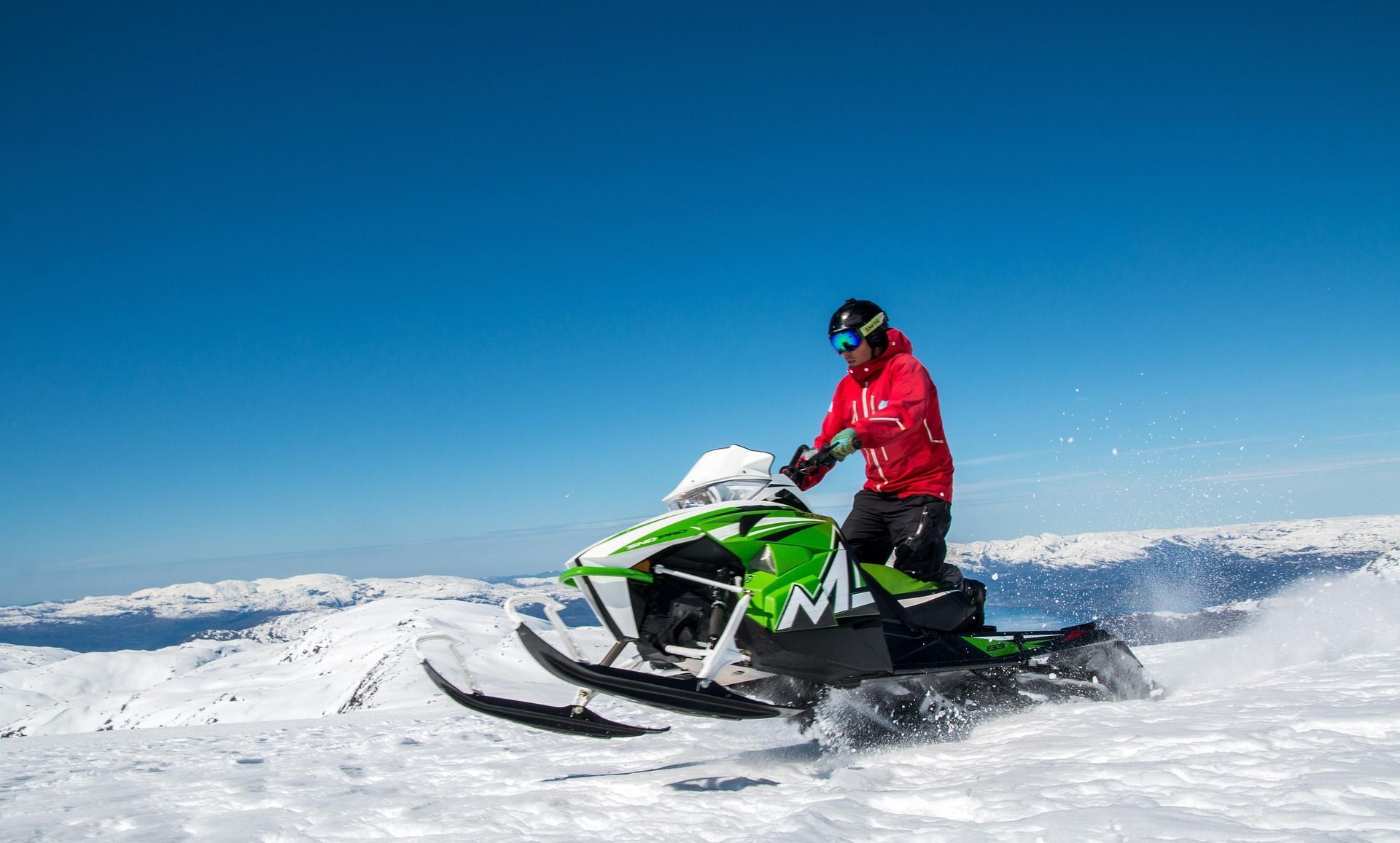 Una persona con una chaqueta roja conduce una moto de nieve verde por una cordillera nevada bajo un cielo azul claro.