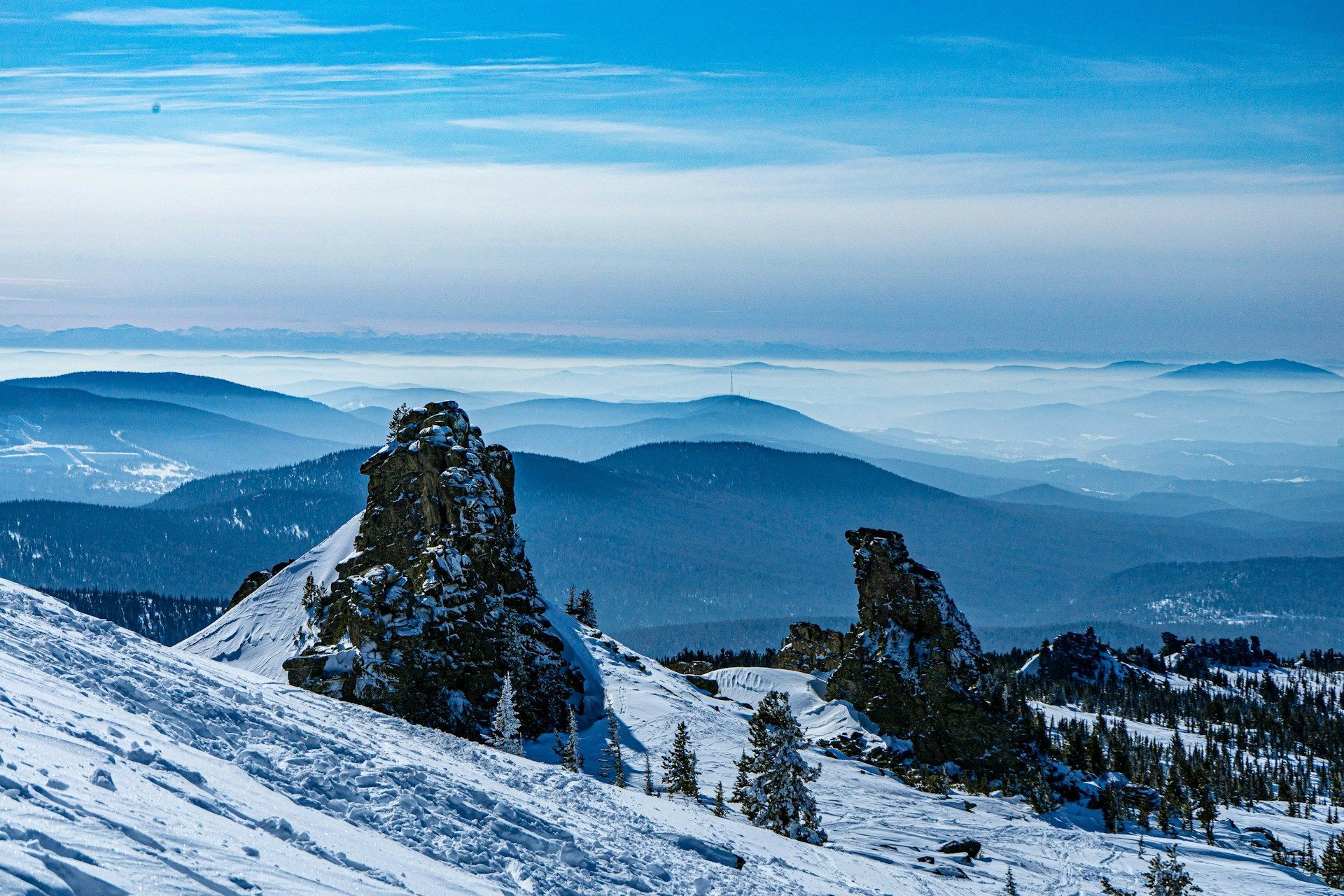 Formazioni rocciose frastagliate sporgono da un fianco di montagna innevato, affacciandosi su strati di montagne azzurre e sfumate che si estendono all'orizzonte sotto un cielo luminoso.