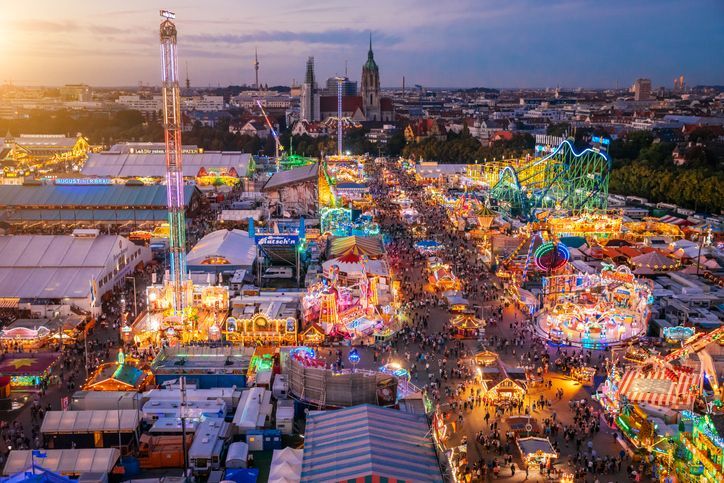 An aerial view of a crowded amusement park at dusk, with colorful lights illuminating rides and stalls against a city skyline.